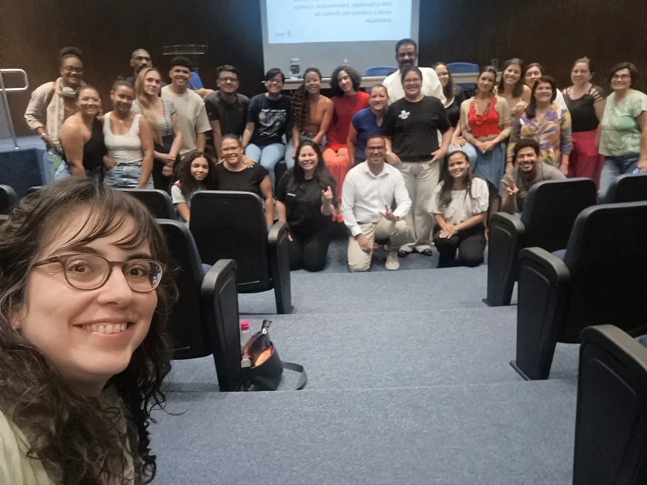 Selfie no auditório, com uma participante em primeiro plano e o grupo da aula sobre Ética e Trabalho Profissional reunido ao fundo.